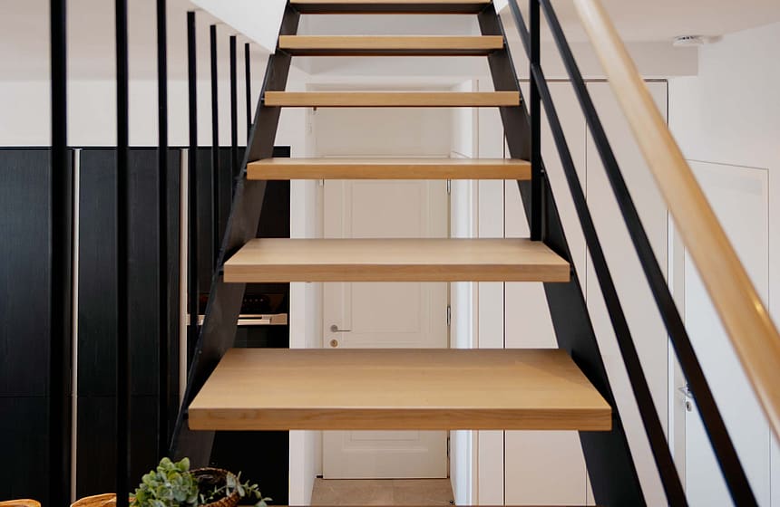A modern, open-tread staircase featuring light oak steps and a sleek black metal railing, ascending toward a bright landing.
