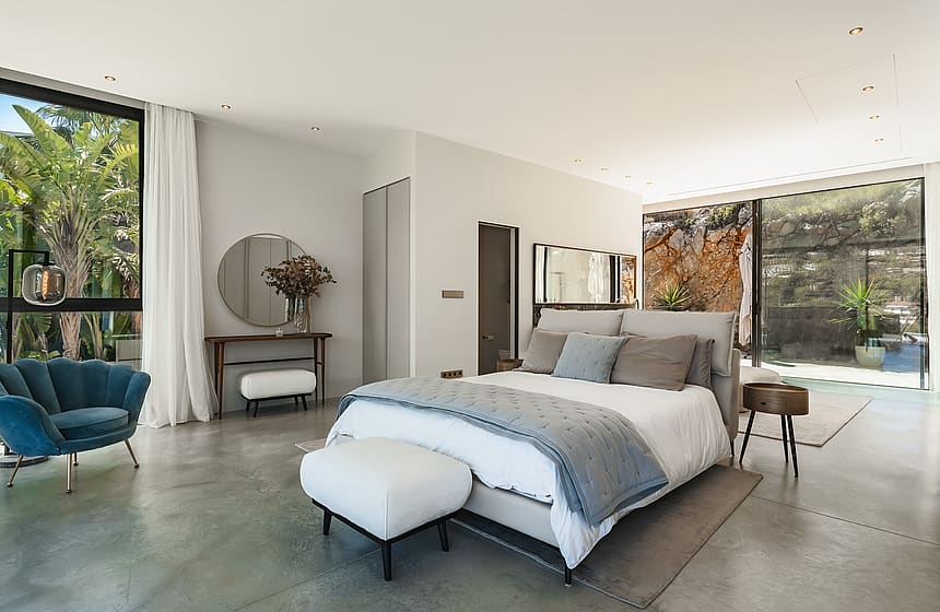 Modern master bedroom with polished concrete floors, floor-to-ceiling windows, a blue velvet chair, and grey bedding.