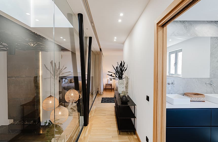 A bright hallway with light wood flooring leading to a master bathroom with a navy vanity, separated by a glass wall.