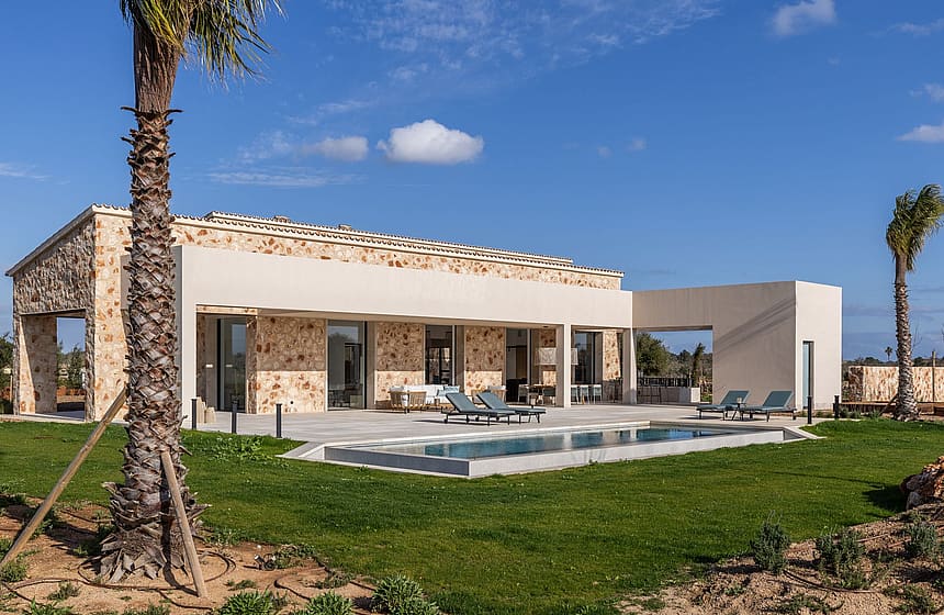 Exterior view of a single-story stone finca in Santanyí with a swimming pool, lawn, and pergola-covered terrace.
