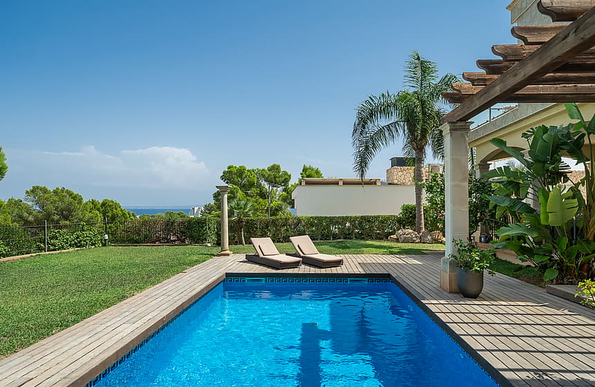 Rectangular swimming pool with blue water next to a timber deck, two sun loungers, and a shaded pergola in a sunny garden.