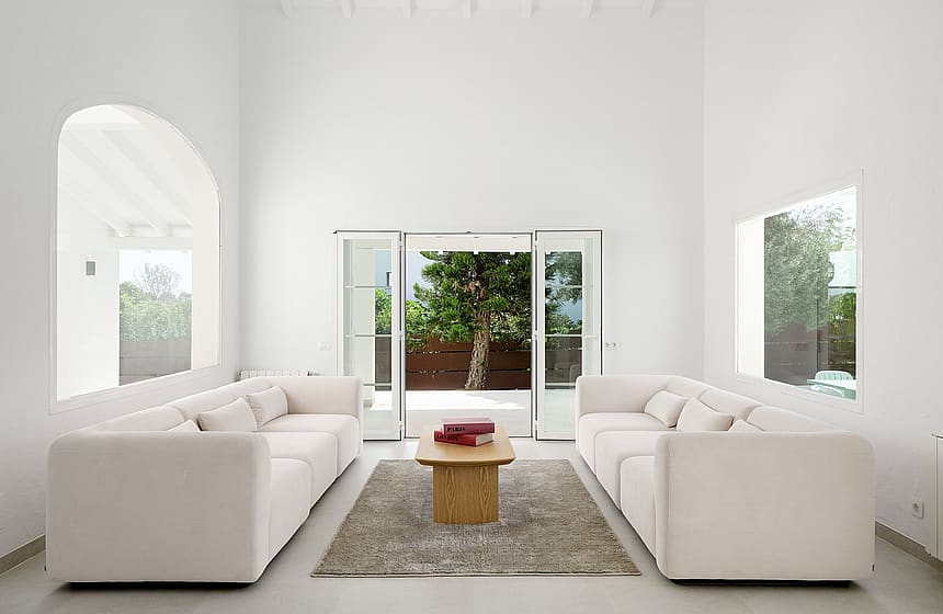 Salón blanco de doble altura con ventana arqueada, dos sofás color crema y puertas acristaladas hacia el jardín