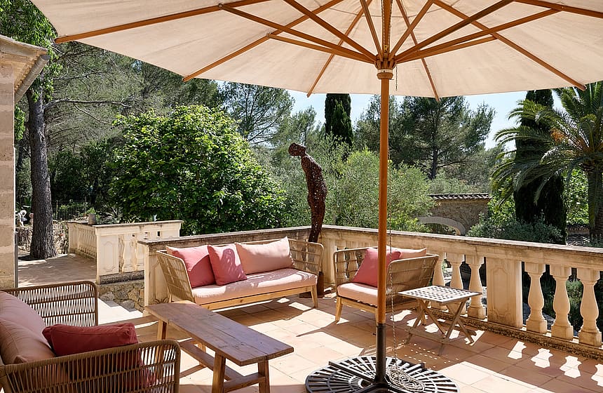 A stone terrace with a balustrade, wicker lounge seating with pink cushions, and a large sun umbrella overlooking trees.