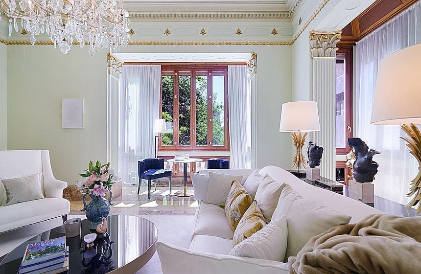 A formal living room with off-white sofas, a crystal chandelier, and ornate crown molding looking toward a windowed alcove.