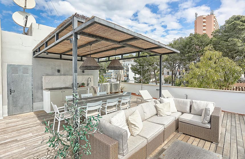 A rooftop terrace featuring a wicker sectional sofa, dining area, and outdoor kitchen under a wooden pergola.