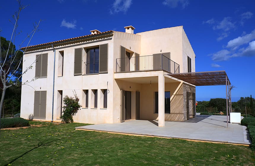 Exterior view of a cream-colored two-story finca with traditional wooden shutters, a shaded terrace, and a grassy garden.