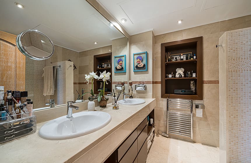 A master bathroom featuring a long stone-topped vanity with double sinks, a large wall-to-wall mirror, and recessed wall shelving.