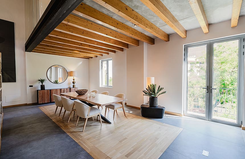 Dining area with wooden table, light-colored chairs, and exposed ceiling beams. Glass doors open to greenery.
