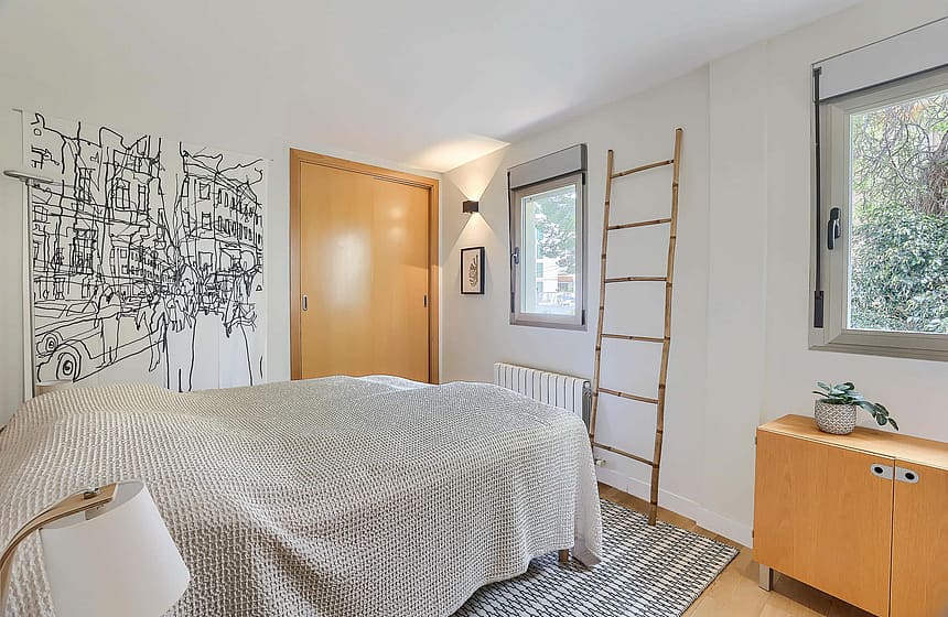 A minimalist bedroom featuring light wood closet doors, white walls, a patterned throw, and a decorative wooden ladder.