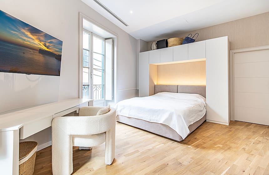 Modern bedroom with a white bed frame surrounded by built-in white cabinets, light oak wood flooring, and a textured white chair.