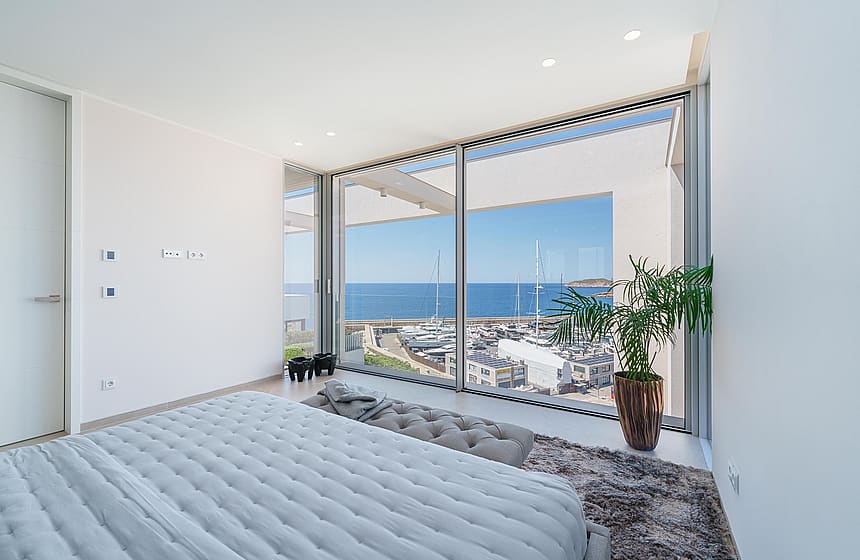 Contemporary bedroom with a quilted grey bed, full-height sliding glass walls, and a view of Port Adriano harbor.