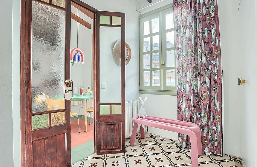 A bright indoor space with patterned floor tiles, a pink bench, a window with floral curtains, and a wooden glass-paneled door leading to another room.