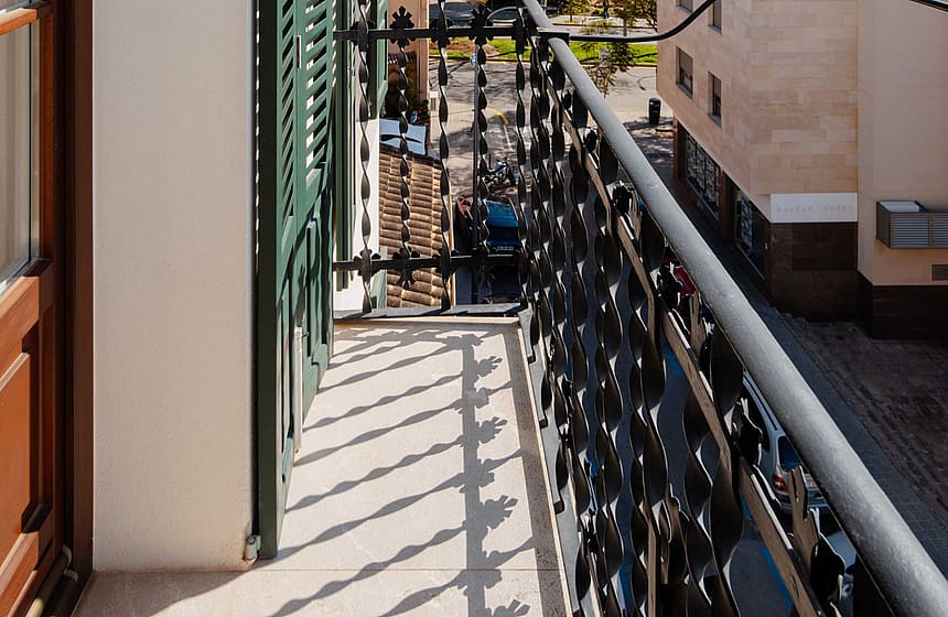 A narrow residential balcony featuring black decorative wrought iron railings and green shuttered doors overlooking a street.