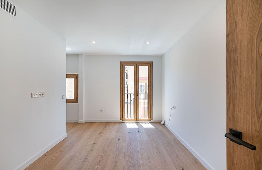 A bright, empty bedroom with light oak wood floors, white walls, and a wooden framed glass door leading to a small balcony.