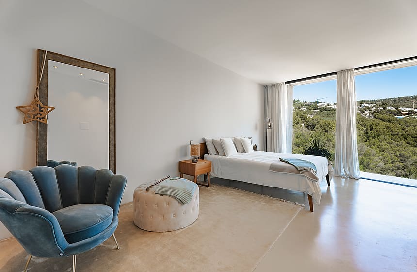 Minimalist bedroom showing floor-to-ceiling windows with green mountain views, a blue shell chair, and light stone flooring.
