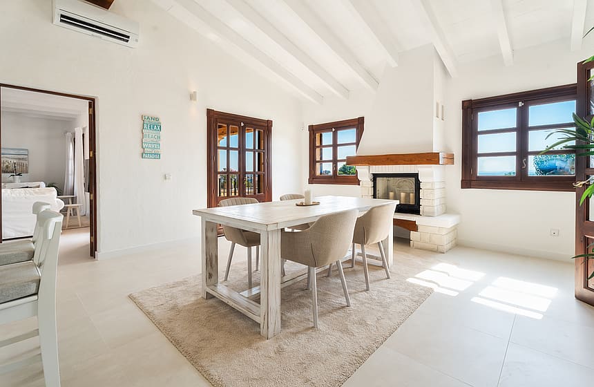 Zona de comedor con mesa de madera, sillas de mimbre y chimenea de piedra sobre muros blancos.