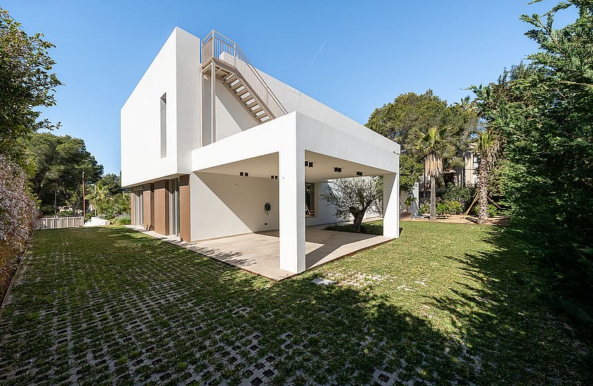 Side view of a white, two-story modern villa with a covered terrace, external metal staircase, and a green grass garden.