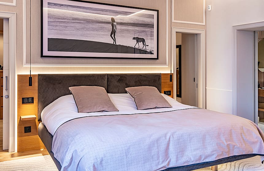 A modern master bedroom featuring a grey upholstered headboard, wooden paneling, and an oversized framed art piece above.