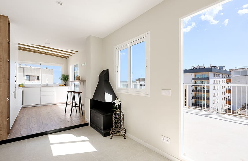 Bright open-plan living space featuring a black conical fireplace, white walls, and a glass door opening to a sunlit balcony.