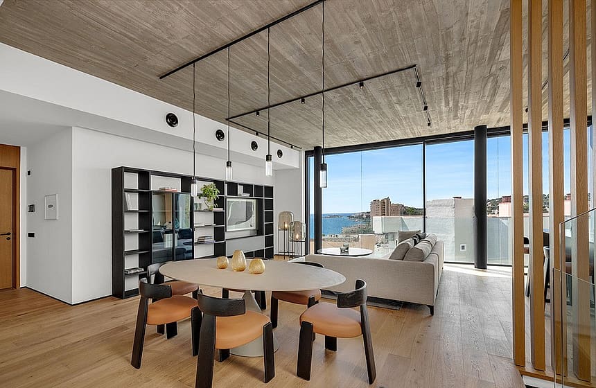 Modern open-plan living and dining room with concrete ceiling, wooden floors, and floor-to-ceiling windows overlooking the sea.