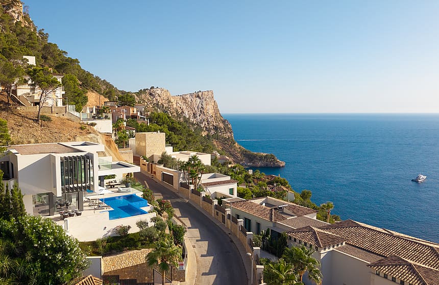 Aerial view of a white cliffside villa with a blue swimming pool, situated along a coastal road overlooking the Mediterranean.