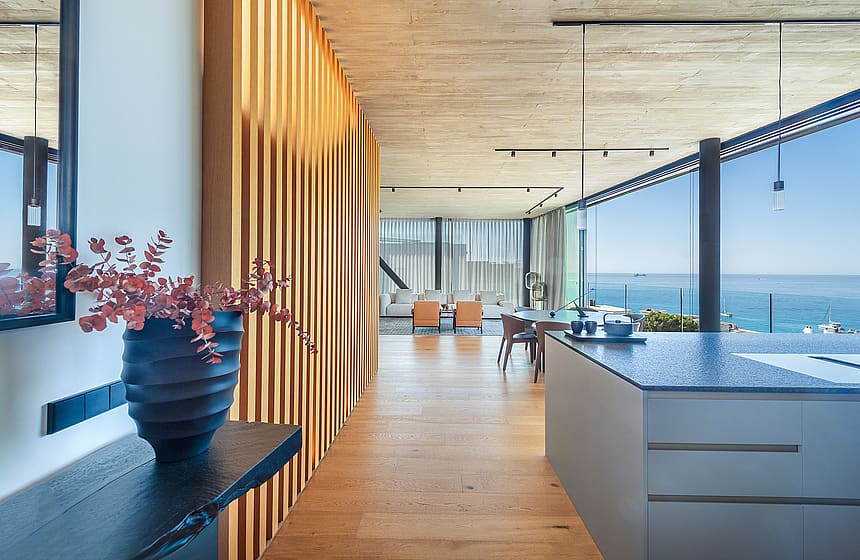 Interior of a modern kitchen showing a textured wood slat wall divider, dark kitchen island, and open-plan living area with horizon sea views.