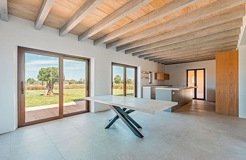 Dining area with a stone table and dark metal legs, exposed wooden ceiling beams, and large glass doors overlooking a rural landscape.