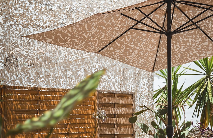 A shaded outdoor terrace with a dining table and rattan chairs, featuring a camouflage net canopy and wooden pallet screens.