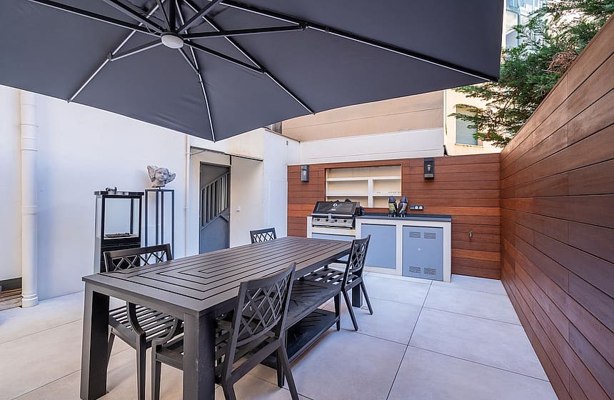A private outdoor terrace in Palma featuring a charcoal grey dining set under a large umbrella, adjacent to a wooden-paneled summer kitchen.