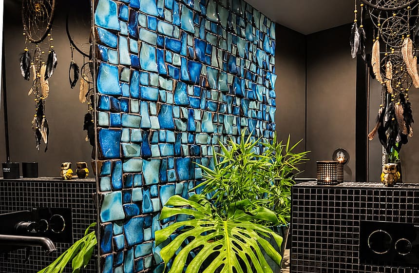 Interior of a bathroom featuring a bright blue mosaic tile wall, a black vessel sink, and a large monstera plant.