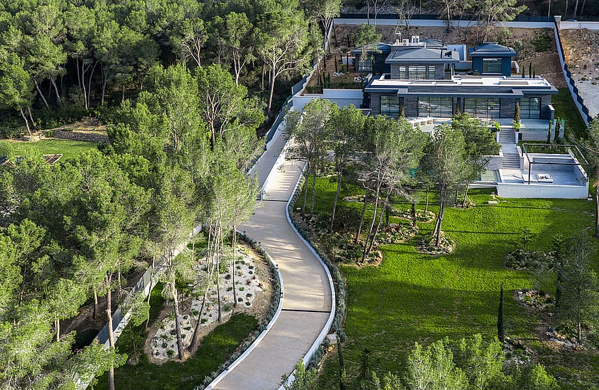 Aerial view of a modern villa with a flat roof, surrounded by gardens, pine trees, and a winding driveway.