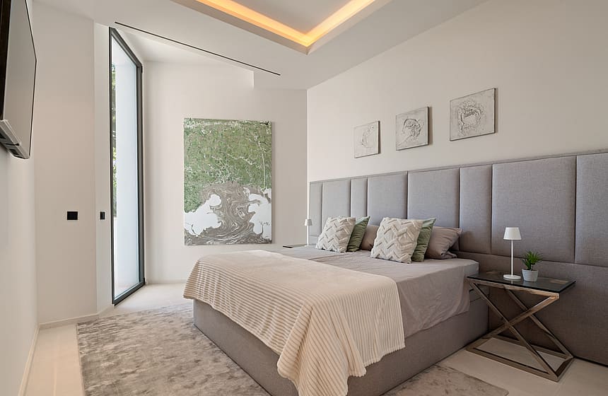 A minimalist bedroom featuring a king-sized bed with a tall grey upholstered headboard, a textured area rug, and cove lighting.