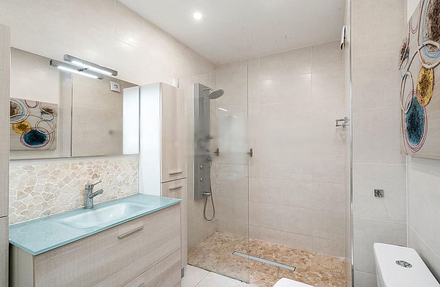 A contemporary bathroom featuring a light-toned wooden vanity, glass sink, and a walk-in shower with textured stone floor tiles.