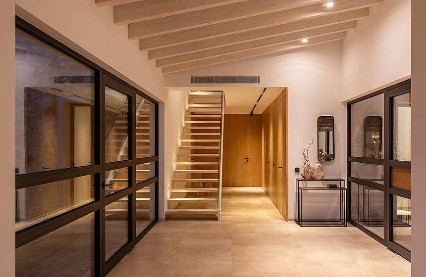 Interior entrance hall of a modern finca featuring a floating wooden staircase, light-toned stone flooring, and timber ceiling beams.