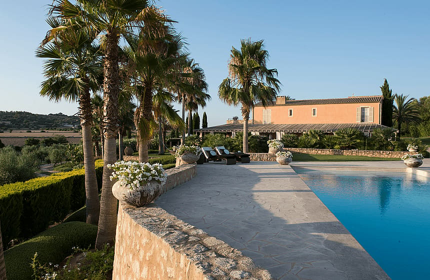 Outdoor swimming pool with stone terrace, palm trees, and terracotta finca building in the background.