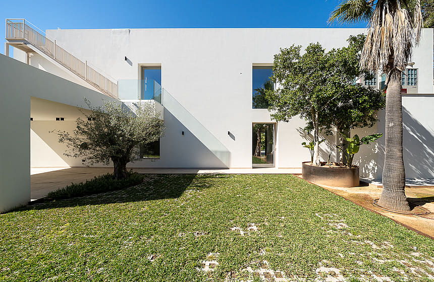 Modern white minimalist villa exterior featuring a flat roof, large rectangular windows, and a manicured green lawn with mature trees.