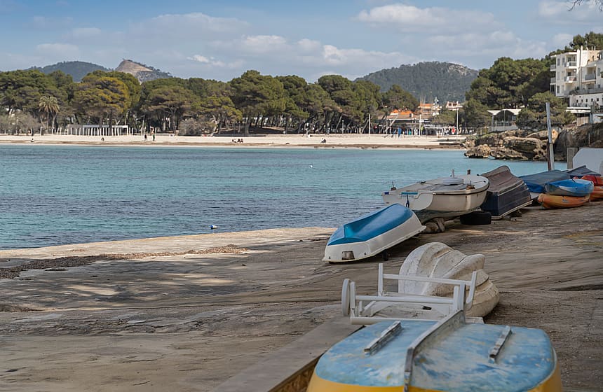 Small boats parked on a stone ramp facing a calm turquoise bay with a sandy beach and dense pine forest in the distance.