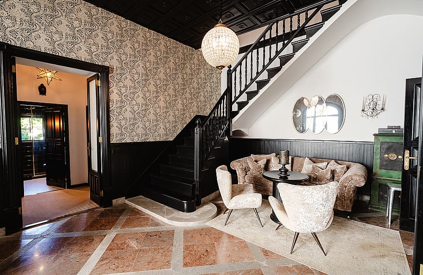 Entrance foyer featuring a dark wood staircase, patterned wallpaper, a crystal pendant light, and a seating area with ornate chairs.