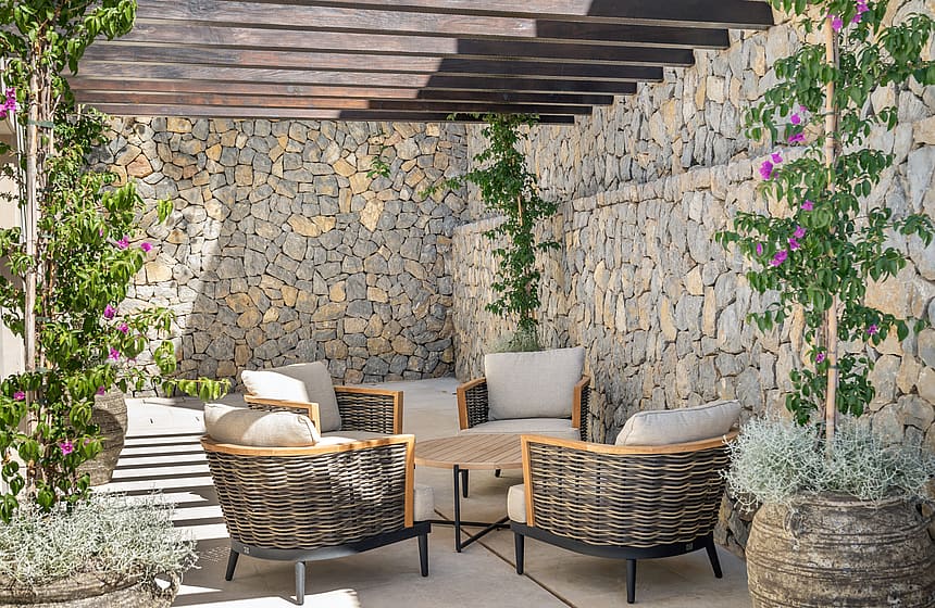 An outdoor lounge featuring woven armchairs set against a textured dry stone wall under a slatted wooden pergola.