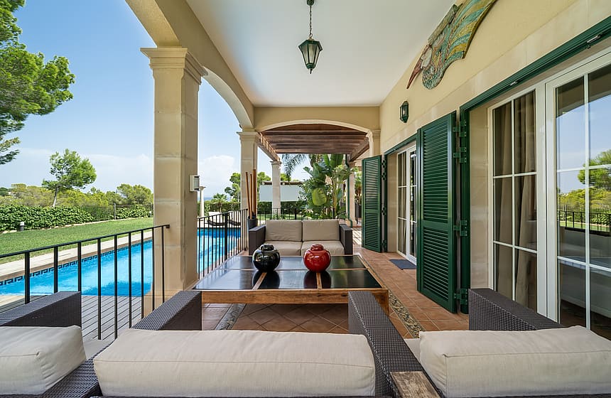 A wide covered terrace with dark rattan furniture and green louvered shutters next to a swimming pool on a sunny day.