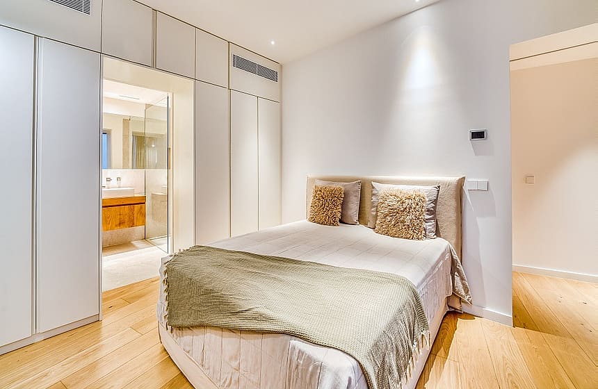 A minimalist bedroom featuring floor-to-ceiling white wardrobes, light oak wood flooring, and a bed with textured cushions.