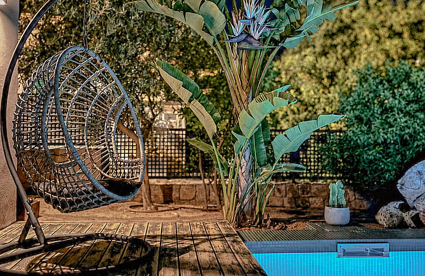 A poolside wooden deck at night with an illuminated swimming pool, a hanging wicker chair, and lush tropical foliage.