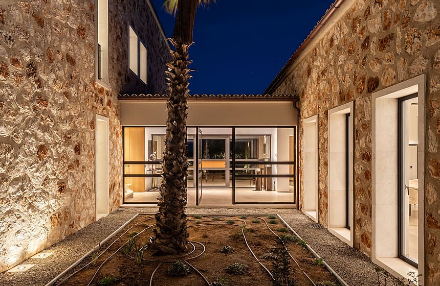 A central courtyard in a stone-clad finca at dusk, featuring a palm tree in a gravel bed and illuminated glass doors.