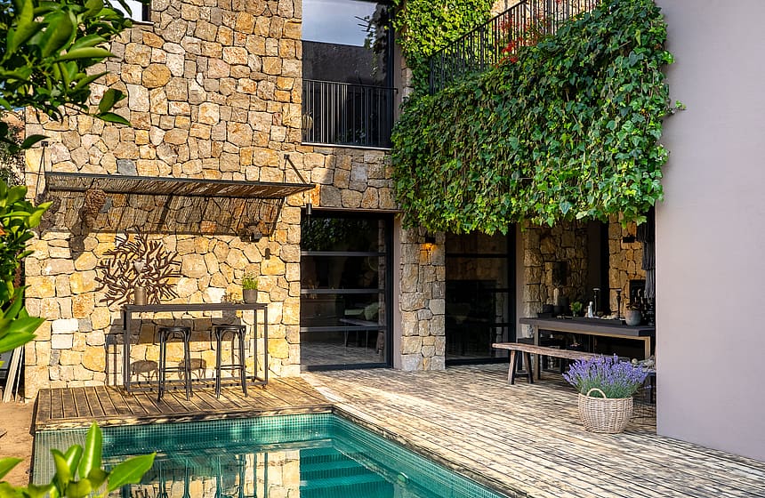 A stone villa facade with a swimming pool, outdoor dining table, and climbing greenery on the terrace wall.