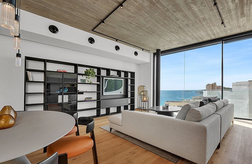 Modern living room featuring a concrete ceiling, oak flooring, large floor-to-ceiling windows overlooking the sea, and a minimalist black shelving unit.