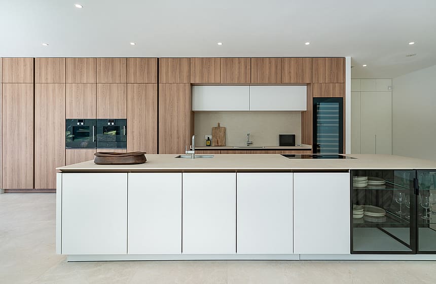 White handleless Siematic kitchen island with stone worktop, integrated sink and glass-fronted crockery display, floor-to-ceiling oak-veneer cabinetry and Gaggenau ovens behind