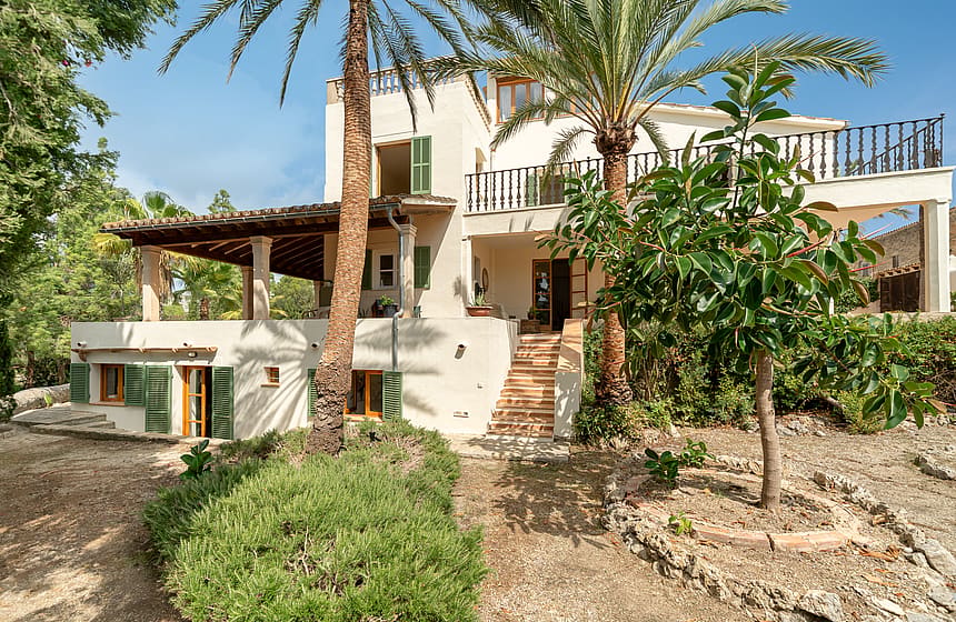 Multi-level Mediterranean villa with cream stucco walls, green shutters, and stone steps framed by palm trees.