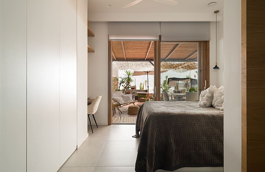 A minimalist bedroom featuring floor-to-ceiling white wardrobes, beige stone floors, and glass doors opening to a furnished terrace.