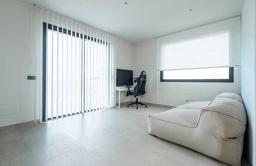 A minimalist room featuring a white desk, a black ergonomic chair, and a large beige floor cushion, with vertical blinds on a sliding glass door and polished concrete floors.