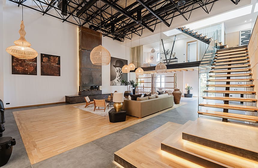 A spacious apartment interior with high ceilings, exposed metal roof trusses, a floating staircase, and neutral-toned furniture.